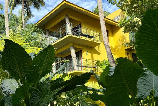 Yellow villa at Waterwoods Havelock surrounded by lush tropical greenery and tall coconut trees under a blue sky.
